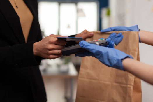 Closeup of businesswoman paying takeaway food order with credit card using POS contactless service during takeout lunchtime. Delivery man with medical face mask and gloves giving fresh food meal.