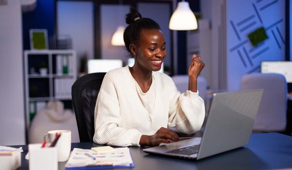 Happy african freelancer receving good news on laptop working overtime in start up company office. Enthusiastic winner employee using modern technology celebrate online win success reading great data.