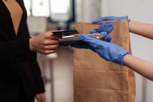 Closeup of entrepreneur woman paying takeaway food meal order with credit card using POS contactless terminal working in startup office. Delivery man bringing lunch box at workplace