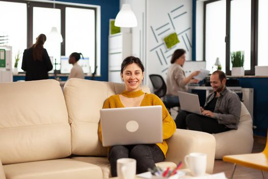 Businesswoman sitting on couch holding laptop, smiling at camera while diverse colleagues working in background. Multiethnic coworkers talking about start up financial company in modern business office