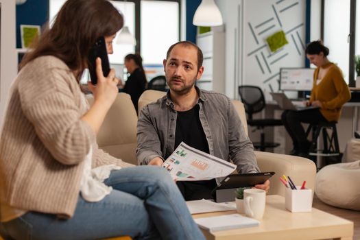 Employee taking at smartphone with business partner sitting on couch looking at documents. Woman discussing on phone while multiethnic coworkers planning financial project working in open plan office