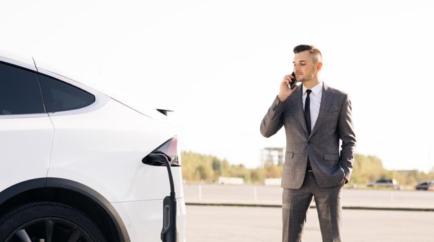 Businessman using smartphone and leans on white luxurious electric car that is charging. Plug charging an electrical car. Luxury electrical car recharging.