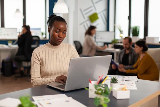 Portrait of authentic african business woman reading email on laptop sitting at desk in busy start up office while diverse team analyzes statistics data. Multiethnic team working at new project