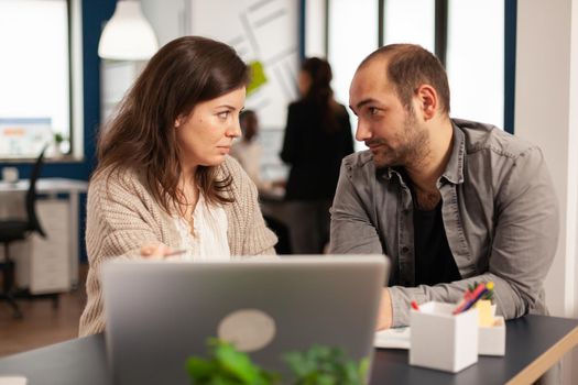 Business man looking at woman colleague and talking about business plan using laptop sitting at table in creative cozy office concentrated on job, while diverse team working in background.