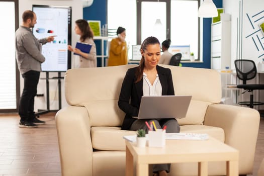 Enthusiastic manager reading report, typing on laptop sitting on couch in busy start up office while diverse team analyzes statistics data in background. Multiethnic team talking about project