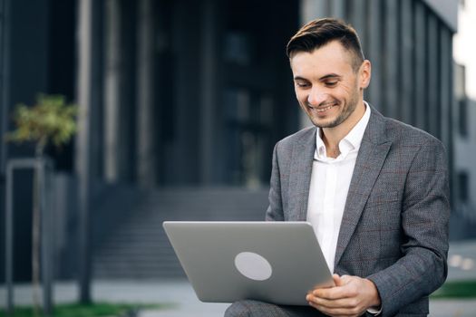 Close up of confident businessman working on laptop outdoors. Stylish successful entrepreneur talking on video conference on laptop. Modern businessman working with laptop on the bench outdoors.