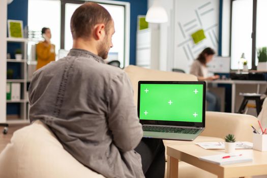 Business team sitting on couch analysing financial statistics, holding laptop with green screen while diverse team working on background. Multiethnic coworkers planning project on chroma key display