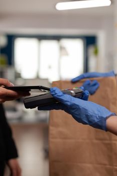 Delivery guy wearing protective gloves receiving paymant from businesswoman using smartphone nfc, technology over pos terminal service for delicious tasty meal lunch in paper bag.