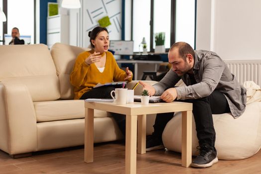 Stressed busineswoman arguing, yealing at man during work hours sitting on the couch, while diverse colleagues working scared in background. Coworkers having conflict because of mistakes from project