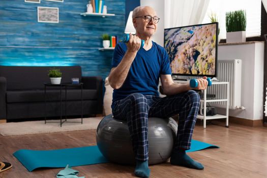 Senior man holding dumbbells doing arms exercise streching body muscles training muscular resistance. Retirement caucasian male sitting on swiss ball in living room doing fitness workout