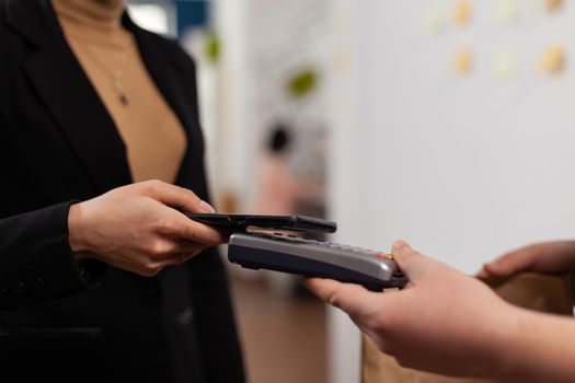 Close up of business woman paying delivery guy from frood service, using smartphone nfc technology. Entrepreneur recieving tasty takeaway lunch in paper bag.