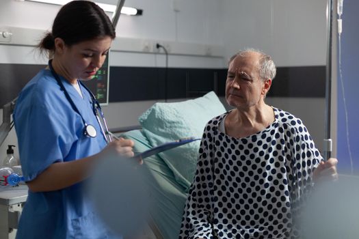 Senior sick man in hospital room discussing with nurse about treatment during consultation. Medical staff with stethoscope writing notes on clipboard preparing patient for surgery.