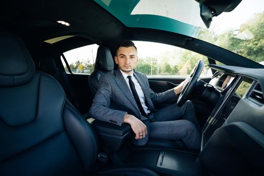 Portrait of male businessman sitting at the wheel of a car. Young cheerful man sitting in modern luxurious car staying in city center