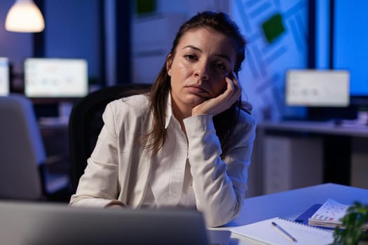 Exhausted businesswoman looking tired in camera sighing resting head in palm late at night in business office. Focused entrepreneur using technology network wireless, working at marketing statistics