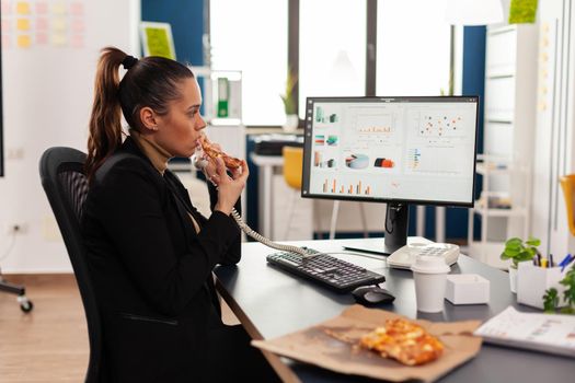 Closeup of businesswoman sitting at desk in front of computer eating pizza slice while talking at landline with remote company manager. Lunch meal break package delivered at startup office.