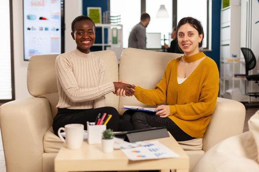 Diverse women looking at camera smiling, shacking hands after signing investment papers contract with confident partner in casual wear. Multiethnic businesswomen making satisfactorily agreement