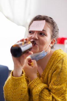 Happy woman with sticky note on forehead playing name game with multi ethnic friends keeping social distancing drinking beer in home living room due to social pandemic. Conceptual image.