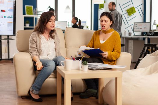 Woman leader yelling at employee sitting on couch in new start up business, upset of bad contract agreement. Women colleagues having big conflict, dispute about mistakes from project documents