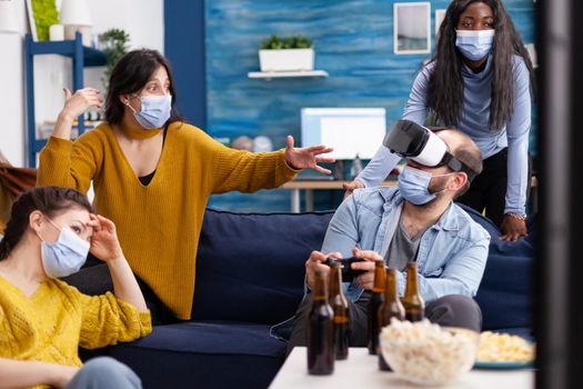 Group of multi ethnic friends having fun playing video games experiencing virtual reality using vr headset keeping social distancing in home living room due to social pandemic.
