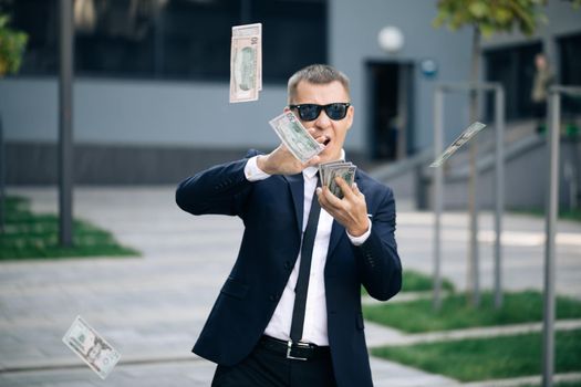 Money rain, falling dollars. Successful business concept. Successful Businessman in a Suit Wear Sunglasses Throwing Money in the Camera Standing in the Street near Office Building.