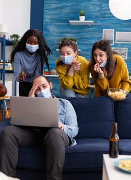 Cheerful group of multi ethnic friends laughing together keeping social distancing and wearing face mask to prevent cooronavirus spread during global outbreak watching video on laptop in living room.