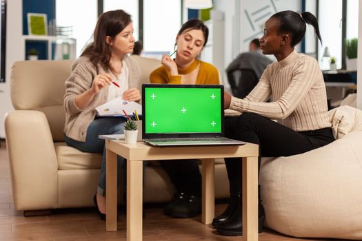 Laptop with chroma key display on table in startup business workplace. African manager discussing talking project statistics with employees on modren office sitting on couch.