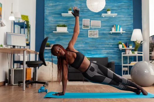 Fit athletic black woman standing in side plank on yoga mat in home living room, doing strenght exercises, training for healthy lifestyle. Full lenght of african american working out.