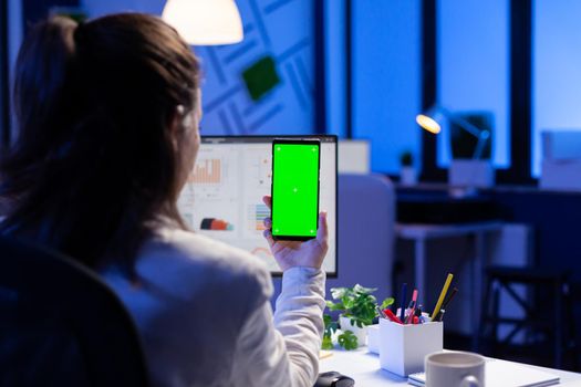 Entrepreneur looking at smartphone with green screen sitting at office desk overworking. Manager project watching desktop monitor display with mockup chroma key during videocall meeting remote
