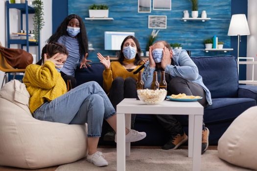 Group of multi ethnic friends watching a horror movie on tv being afraid wearing face mask to prevent infection with covid 19 during global pandemic having fun sitting on couch and