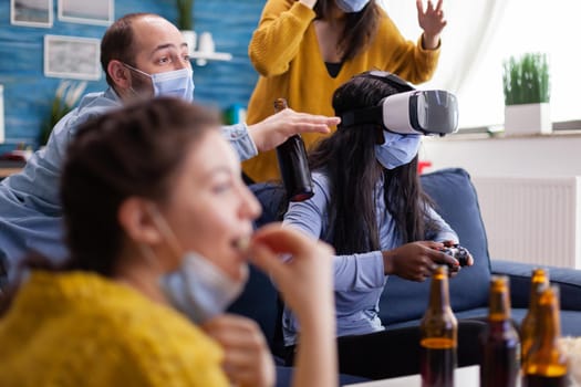 African woman experiencing virtual reality playing video games with vr headset and joystick in home living room while friends are cheering up for her keeping social distancing because of global pandemic.