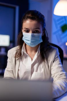 Close up of businesswoman with face mask checking emails late at night in new normal business office before deadline. Taking notes, analysing documents overtime during global pandemic