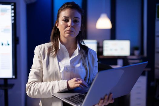 Exhausted entrepreneur woman holding laptop, typing business emails and looking at camera standing in start-up company late at night. Businesswoman using technology network wireless making new project