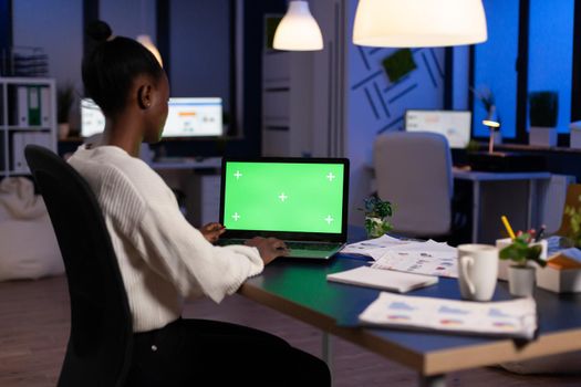Dark skin freelancer working on laptop with green screen display sitting at desk in business office late at night. Businesswoman watching desktop with green mockup, chroma key, working overtime.