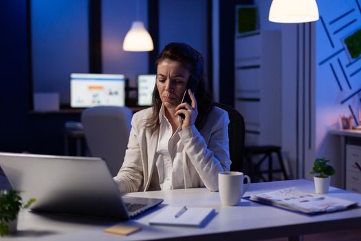 Businesswoman having phone call in start-up office late at night working at marketing project to respect deadline. Exhausted employee checking business paperwork analysing financial profit