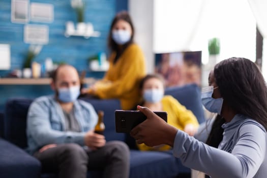 African woman showing video clip to cheerful friends on smartphone in home living room wearing face mask as prevention against the spread and infection of covid19 while hanging out. Conceptual image.