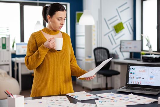 Business woman holding cup of coffee looking at charts on paperwork. Executive entrepreneur, manager leader standing working on documents projects. Successful corporate professional entrepreneur working on online internet statistics