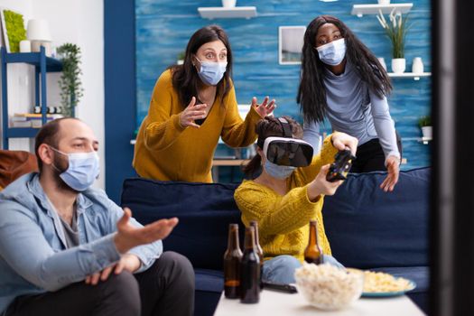 Group of multi ethnic friends having fun playing video games using virtual reality headset and joystick wearing face mask keeping social distancing to avoid coronavirus spread. Conceptual image.