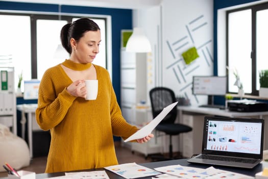 Confident business woman holding documents with statistics enjoying a cup of coffee. Executive entrepreneur, manager leader standing working on documents projects, Successful corporate professional entrepreneur working on online internet