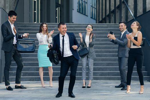 Young Happy Business Manager Wearing a Suit and Tie Dancing from Office Building. Colleagues are Cheering. Diverse and Motivated Business People from Modern Office