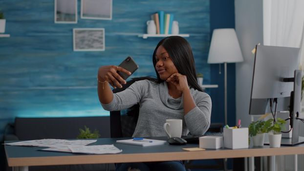 Attractive black student smiling at camera while taking selfie with smartphone sitting at desk table in living room posing on social media. Blogger woman relaxing putting photo on internet