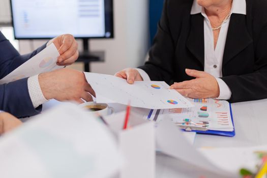 Close up view desk full of papers stats shown on charts and diverse businesspeople. Colleagues working in office at conference table in startup management company.