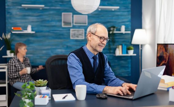 Happy senior man after reading a email with good news sitting at desk. Elderly man entrepreneur in home workplace using portable computer sitting at desk while wife is holding tv remote.
