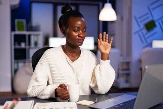 Black businesswoman saying hello during video call sitting at desk, late at nigh doing overtime. Entrepreneur manager working to finish important deadline, having conference discussion.