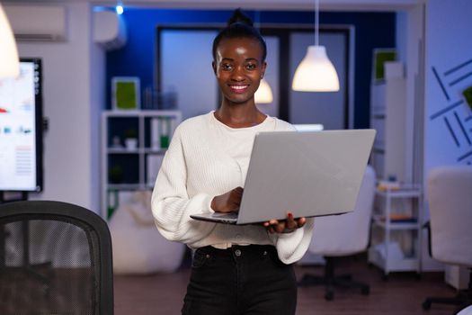 Black business woman looking at camera smiling holding laptop standing near desk in start-up company late at night. Focused employee using technology network wireless doing overtime for job.