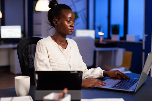 Focused african entrepreneur writing on laptop late at night to finish deadline. Busy multitasking employee analysing financial statistics overworking writing, searching.