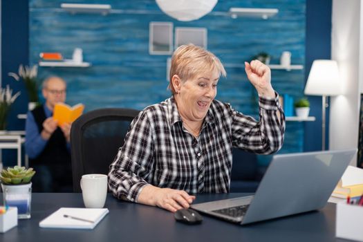 Excited mature woman celebrating online purchase cheerful elderly woman while using laptop computer in living room while husband is reading a book sitting on sofa.