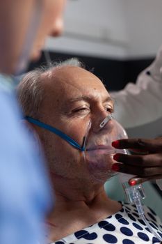Side view of senior patient breathing assisted by respiratory tube in hospital intesive care. African doctor and nurse helping old man to breath sing face mask.