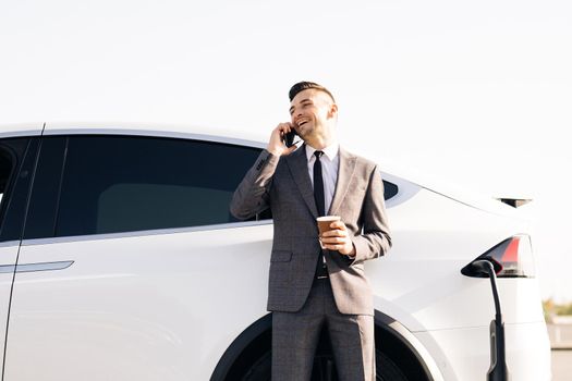 Businessman is talking on his Smart Phone while his electric car is charging on background. Side view of white luxury car. Environmentally conscious male charging electric vehicle.