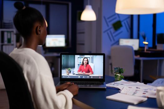 African freelancer working remotely discussing with woman partner online sitting in front of laptop in start-up office talking during virtual meeting at midnight, doing overtime.
