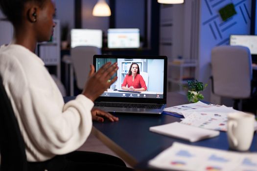 Dark skin manager woman talking with coworker during video conference at midnight from business office planning financial strategy. Worker using wireless headphones discussing at virtual meeting.